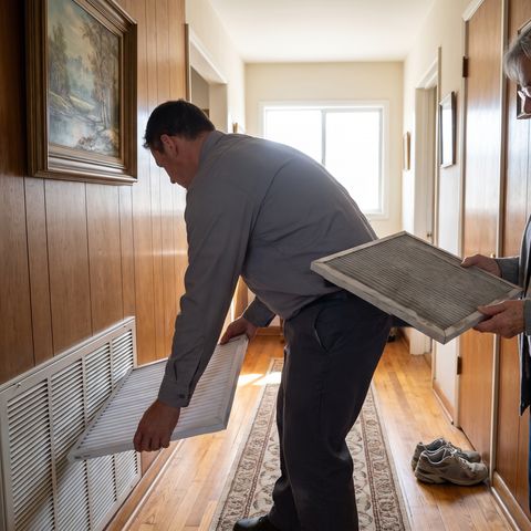 A technician fits a new filter in a hallway return grille while the homeowner holds the old one